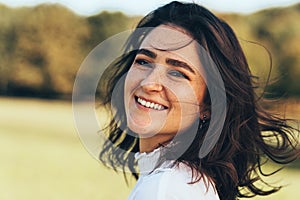 Close-up outdoor portrait of happy beautiful smiling young woman with windy hair and freckles, enjoying the warm weather, posing