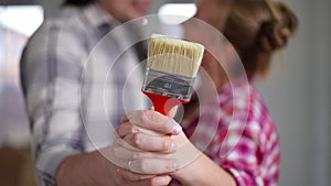 Close-up paint brush in hands of Caucasian man and woman hugging at background. Blurred unrecognizable happy couple in