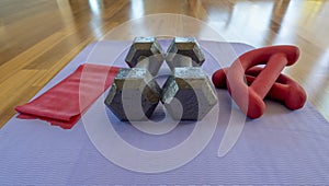 Close up of a pair of dumbbells, theraband exercise bands, and a yoga mat on hardwood floor