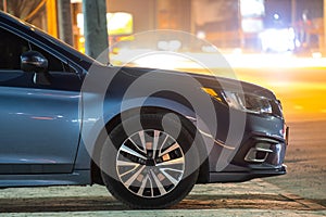 Close up of parked car on roadside at night with blurred view of traffic lights of moving vehicles on city street