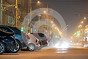 Close up of parked cars on roadside at night with blurred view of traffic lights of moving vehicles on city street