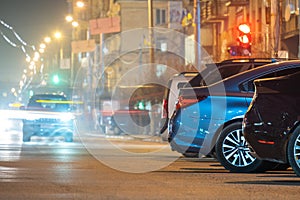 Close up of parked cars on roadside at night with blurred view of traffic lights of moving vehicles on city street