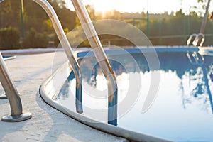 Close-up of a part of swimming pool with a stainless steel ladder and blue water on sunset. Summer vacation, holidays, relax