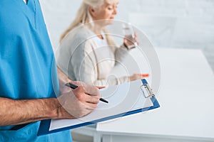 close-up partial view of caregiver writing on clipboard while senior woman