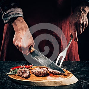 Chef in apron with meat fork and knife slicing gourmet grilled steaks with rosemary and chili pepper on wooden board