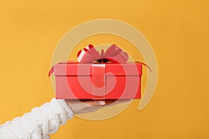 Close-up partial view of man in sweater holding red gift box on yellow background.Gift giving concept