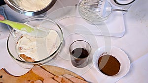 Close-up of pastry chef`s hands smearing ladyfinger biscuits lined in a glass bowl with a layer of sweet cream cheese and whipped