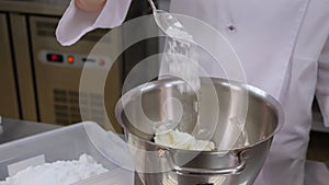 Close-up of a pastry chef spooning powdered sugar into a bowl of buttercream.