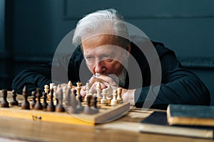 Close-up of pensive gray-haired senior older man thinking game strategy sitting on wooden table with chess board.