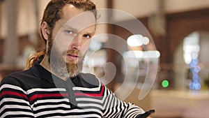 Close-up of pensive male face, Caucasian bearded man with long hair wears striped sweater stands alone looks at screen