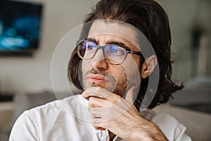 Close up of a pensive mid aged brunette Turkish man face wearing white shirt glasses indoors looking aside
