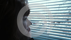 close up of pensive middle-aged woman looking out the window with blinds, looking around, waiting for someone