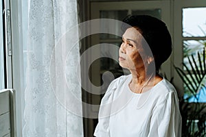 Close up of pensive old woman looking out of window feeling lonely. 70s Asian elderly female pensioner missing children