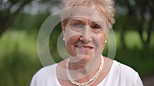 Close-up. Pensive older lady stands on a summer day in a park outdoors. An elderly woman looks at the camera with a