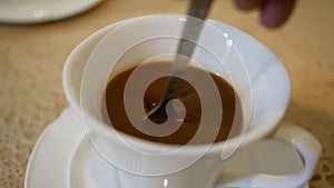 Close-up of person hand stirring coffee with spoon in a white cup on wooden table background.