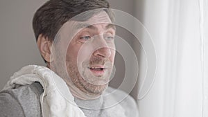 Close-up of perspiring smiling bearded sportsman wiping face with towel and looking out the window. Portrait of