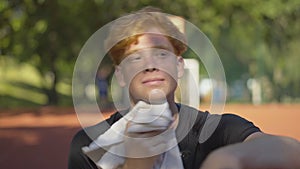 Close-up of perspiring young redhead sportsman wiping face with white towel outdoors. Portrait of confident tired man