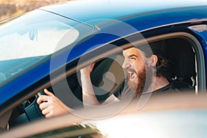 Close up photo of angry driver man with beard screaming while standing in traffic jam