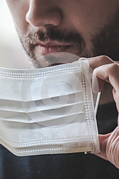 Close up photo of a bearded white man putting on a white medical face mask during coronavirus quarantine. Blurred background,