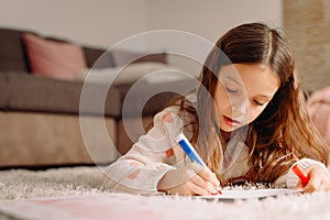 Close up photo of a child with long blond hauir seriosly and attentively writing on the paper with a pen lying down on