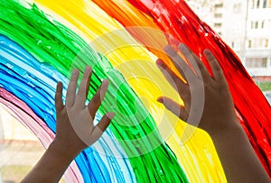 Close-up photo of child`s hands touch painting rainbow on window. Family life background. Image of kids leisure at home