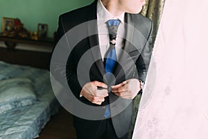 Close-up photo of the elegant groom buttons jacket standing near window