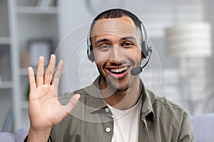 Close-up photo. Face portrait of a smiling young African American man at home wearing a headset talking and greeting the