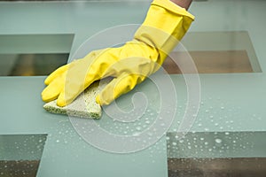 Close-up photo of female hand washing glass table in with cleaning spray and sponge wearing gloves in apartment