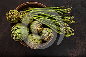 Close up photo of fresh artichoke in the old wooden bowl and Bunch of green asparagus. Top view on dark background