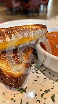 A close-up photo of a grilled cheese sandwich cut in half, revealing melted cheese and a bowl of tomato soup on a white
