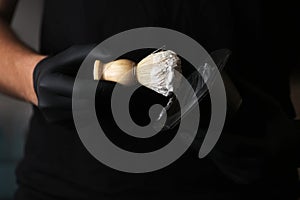 Close up photo of hairstylist hands holding bowl of shaving foam and wooden shaving brush. man He in black glovers is