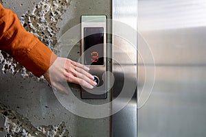 Close-up photo. The hand of a young woman in an orange shirt pressing a metal elevator button with her finger in an