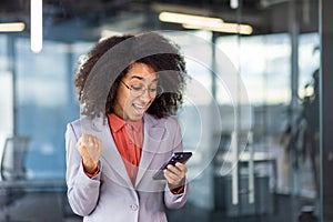 Close-up photo of a happy young African-American woman in a suit standing in the office, looking at the phone screen and