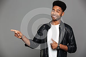 Close-up photo of happy young afro american man in leather jacket pointing with figer while showing thumb up gesture, looking