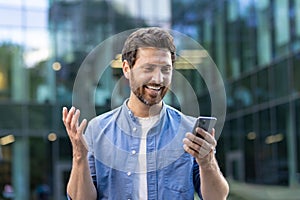 Close-up photo of a happy young man standing on a city street, looking surprised at the mobile phone screen, smilingly photo