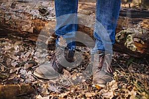 Close up photo of human legs in old leather boots and blue jeans standing on the ground with dried leaves