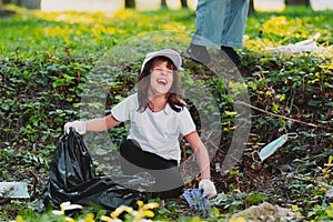 Close up photo of a laughing young girl sitting down on the ground in white gloves holding a garbage bag and picking up