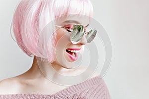 Close-up photo of laughing young girl wears pink wig. Inspired young lady in glasses enjoying photoshoot in colorful