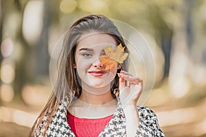 Close-up photo of lovely caucasian girl hiding one eye behind red-yellow autumn maple leaf, posing against blurred