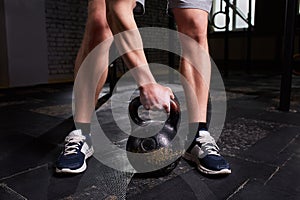 Close-up photo of man`s legs and arm while holding kettlebell on the gym floor against dark background.