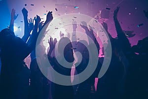 Close up photo of many party people dancing purple lights confetti flying everywhere nightclub event hands raised up