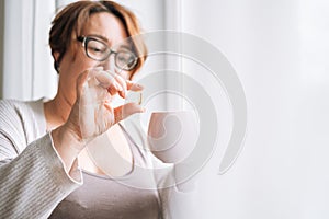 Close up photo of middle aged woman holding omega 3 capsule and mug of water in hands near window at home