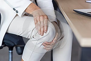 Close-up photo in the office at the desk. The hands of an older woman in a business suit are holding the knee of the leg