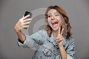 Close-up photo of overjoyed brunette teen girl showing peace gesture while making selfie on mobile phone