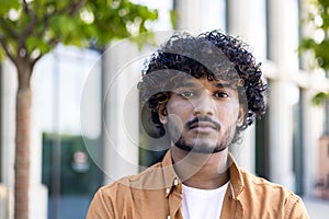 Close-up photo. Portrait, face of a young Indian man in an orange shirt standing outside on the street and looking