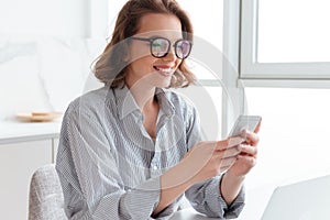 Close-up photo of pretty smiling brunette girl in glasses texting message on smartphone while sitting at kitchen