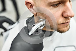 Beard correction. Young handsome man sitting in armchair in barber salon while professional barber trimming beard with