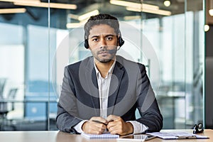 Close-up photo of a serious young Indian male businessman sitting in the office at a desk in front of the camera and