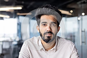 Close-up photo of a serious young Indian man with a beard and a shirt working in the office, sitting and looking