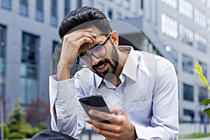 Close-up photo of shocked and upset young Indian man sitting outside holding head with hand and looking at phone screen